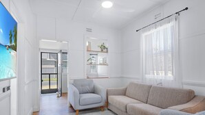 Una sala de estar moderna con un sillón gris, sofá color beige y reposapiés estampado. Se aprecia un suelo de madera y paredes blancas, con una puerta que da a un balcón.