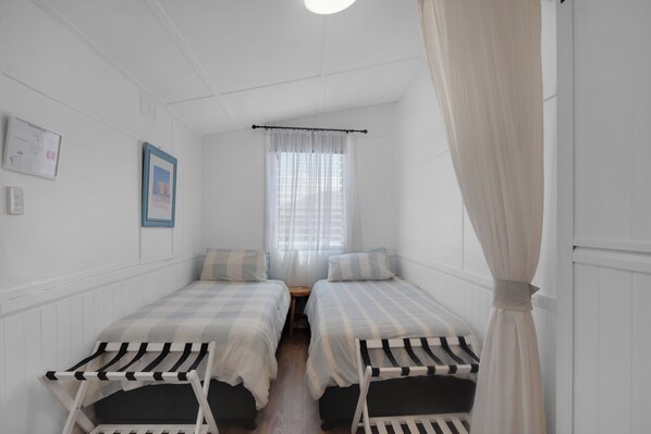 A room with two single beds, a window with sheer curtains, a small table, and a wall-mounted light fixture.