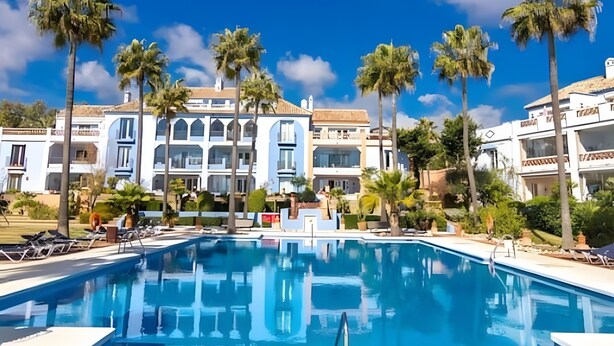 Una piscina con sillas de descanso, palmeras y un edificio grande con varios balcones.