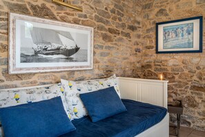 A stone-walled room with a bed, two pillows, a framed photograph of a sailboat, and a framed painting.