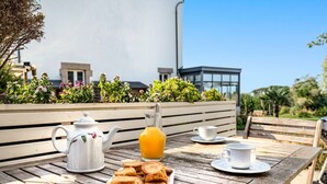 Ein Holztisch mit Teekanne, Orangensaftglas, Teller mit Keksen und einer Tasse auf einer Terrasse mit Blick auf Gebäude und Grünflächen.