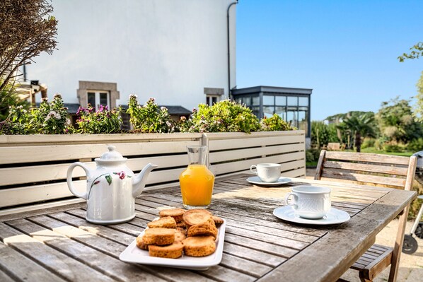 A wooden table with a teapot, a glass of orange juice, a plate of cookies, and a cup on a terrace with a view of buildings and greenery.