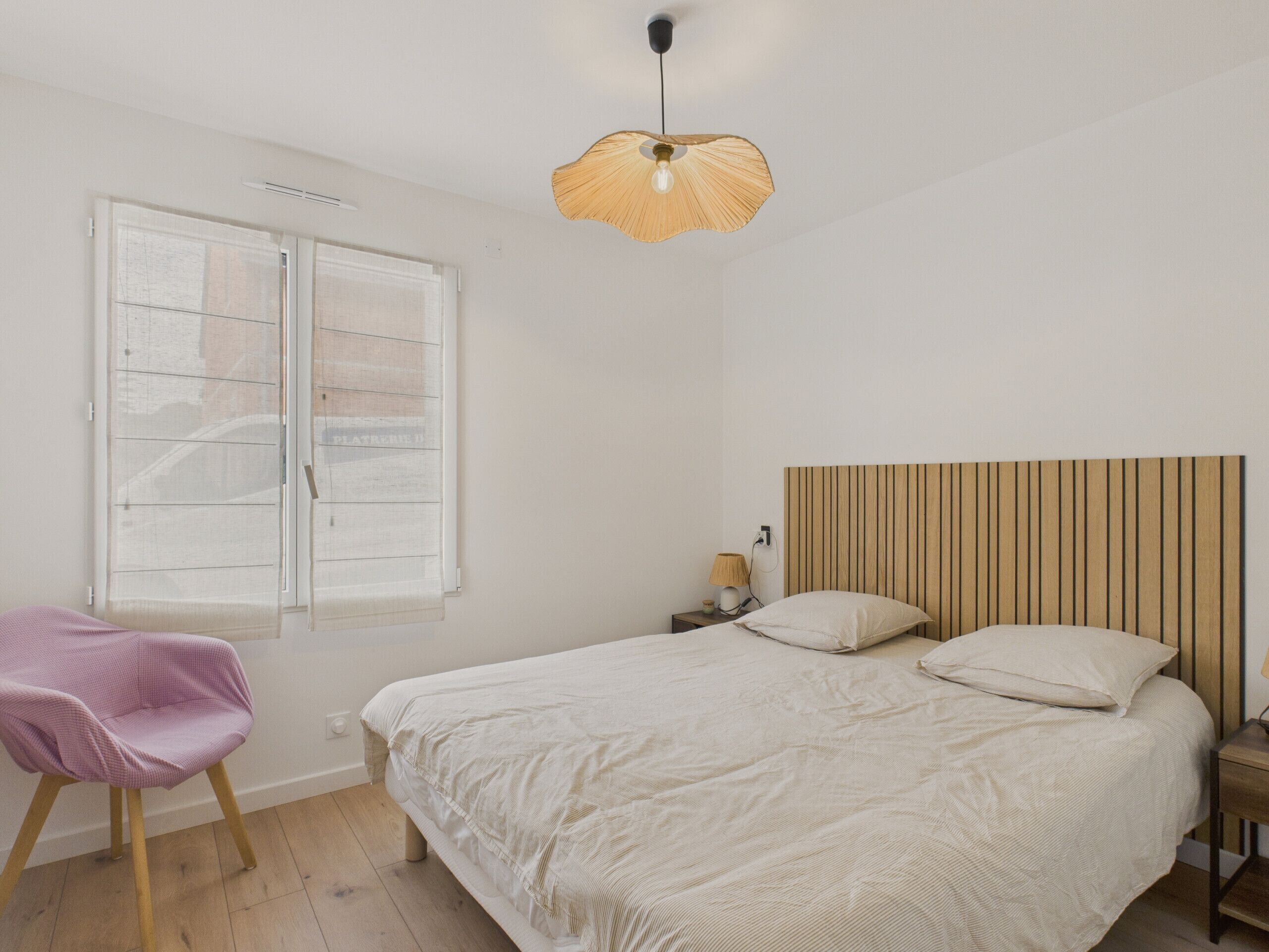 A bedroom with a bed, a chair, a window with blinds, a bedside table with a lamp, and a hanging light fixture.