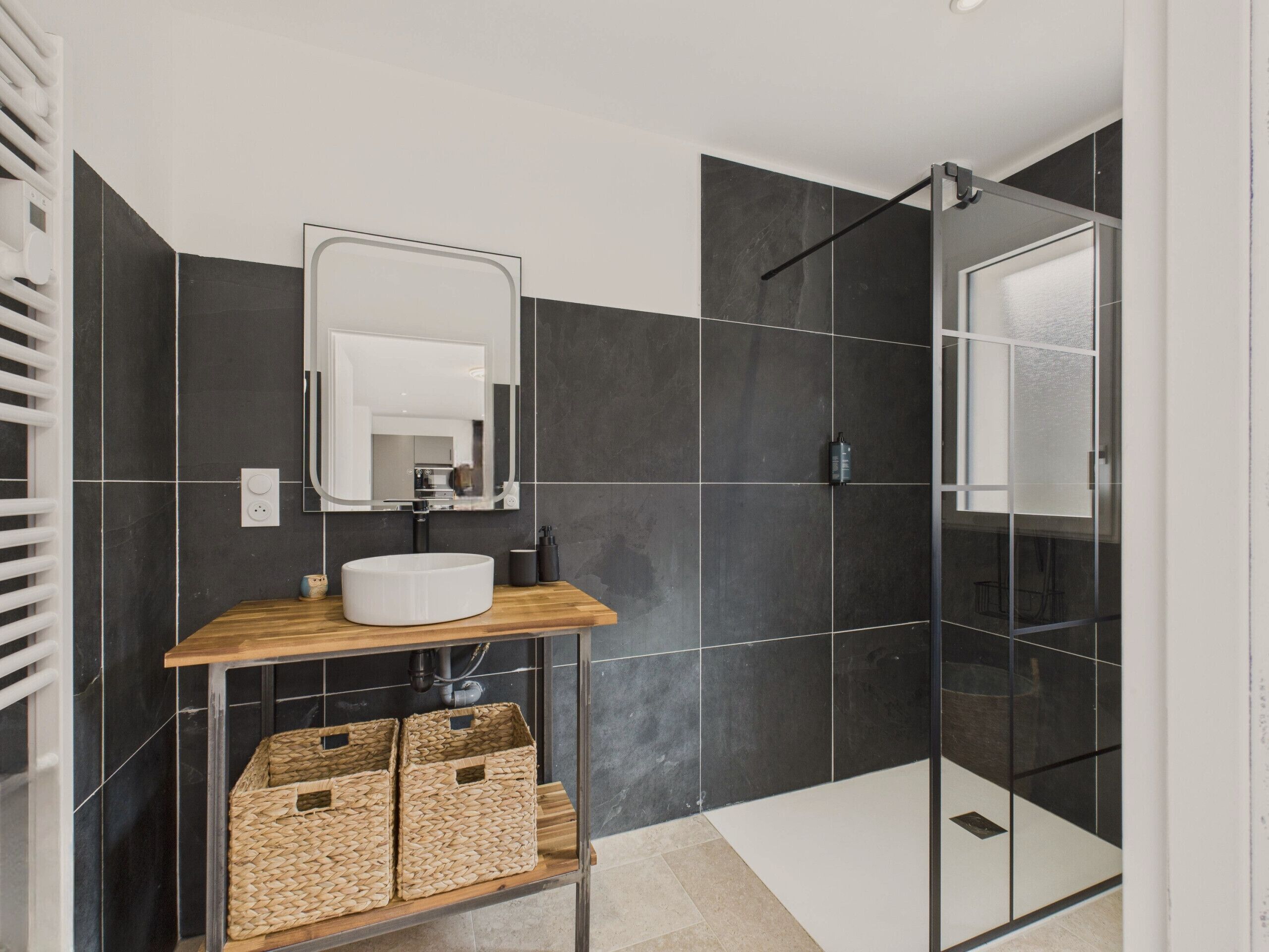 A modern bathroom with a white sink, dark tiled walls, a glass shower enclosure, and a wooden vanity.