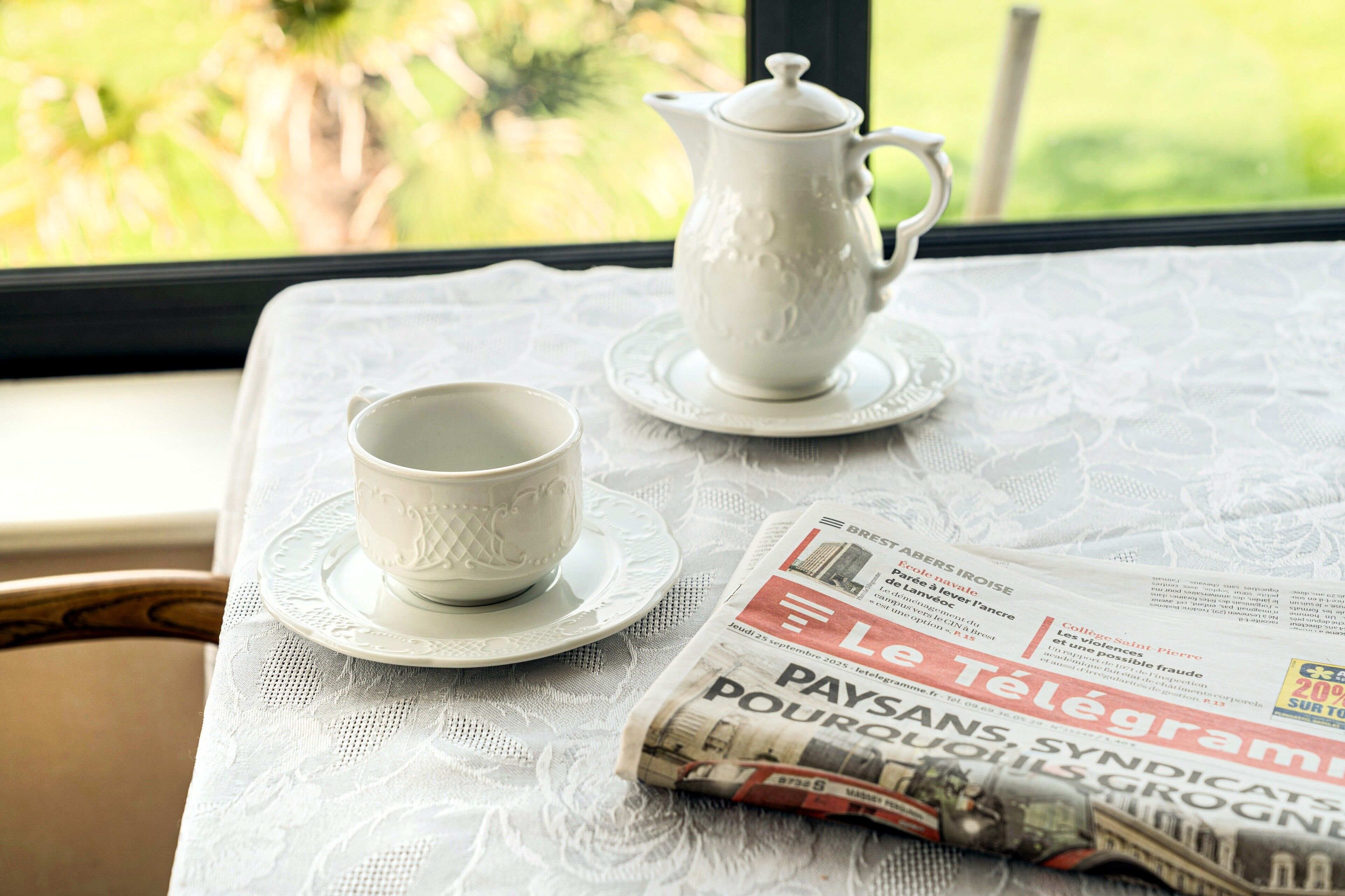 A table with a lace tablecloth, a white teapot, a white cup, and a newspaper.
