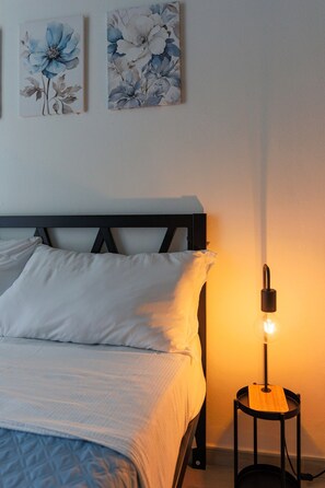 Et soveværelse med seng, puder, et sengebord med lampe og to blomstrede billeder på væggen.