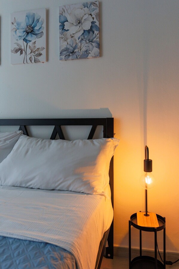 A bedroom with a bed, pillows, a bedside table with a lamp, and two floral wall art pieces.