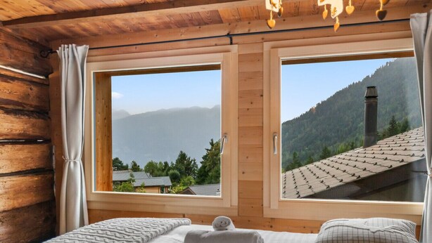 Quarto de cabana de madeira com duas janelas grandes, uma cama com edredom branco e cobertor cinza, e vista para uma paisagem montanhosa.