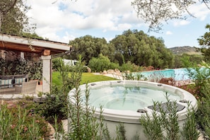 Ein Whirlpool, umgeben von üppiger Vegetation, eine Terrasse mit Gartenmöbeln und Blick auf einen Swimmingpool.