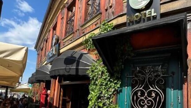 A hotel with a green door and a sign indicating it is a hotel. There are outdoor tables and chairs, and a person is visible inside the building. - Thea Hotel  (Prizren)