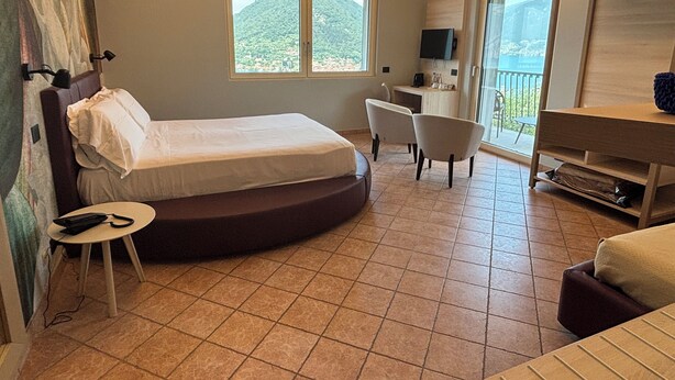 A hotel room with a bed, a desk, a chair, a TV, and a balcony with a view of the mountains. - Pernice77 (Sulzano)