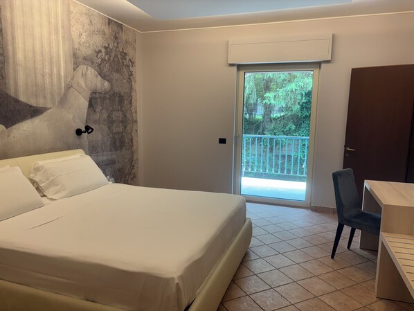 A hotel room with a bed, a desk, a chair, a door, and a balcony with a view of greenery. - Pernice77 (Sulzano)