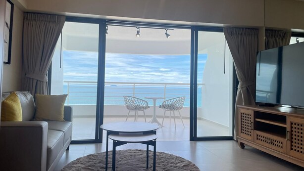 A living room with a sofa, a round table, and a TV on a wooden cabinet. There are two white chairs outside a sliding glass door with a view of the ocean.