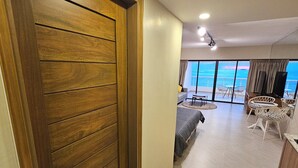 A hotel room with a wooden door, a bed, a seating area with chairs, and a view of the ocean.