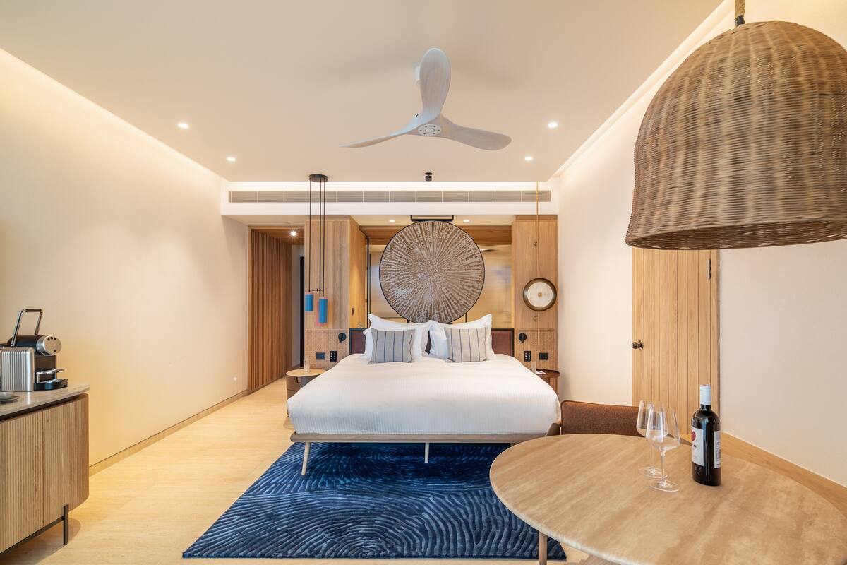 A modern hotel room with a large bed, a dining table with wine glasses, a ceiling fan, and a unique wall art piece.