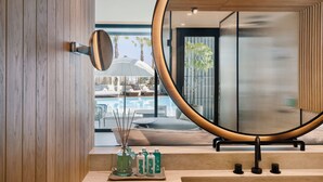 A modern bathroom with a large mirror, wooden paneling, a small sink, and a view of a pool and outdoor seating area. - Kayma By Isrotel Exclusive (Tamar)