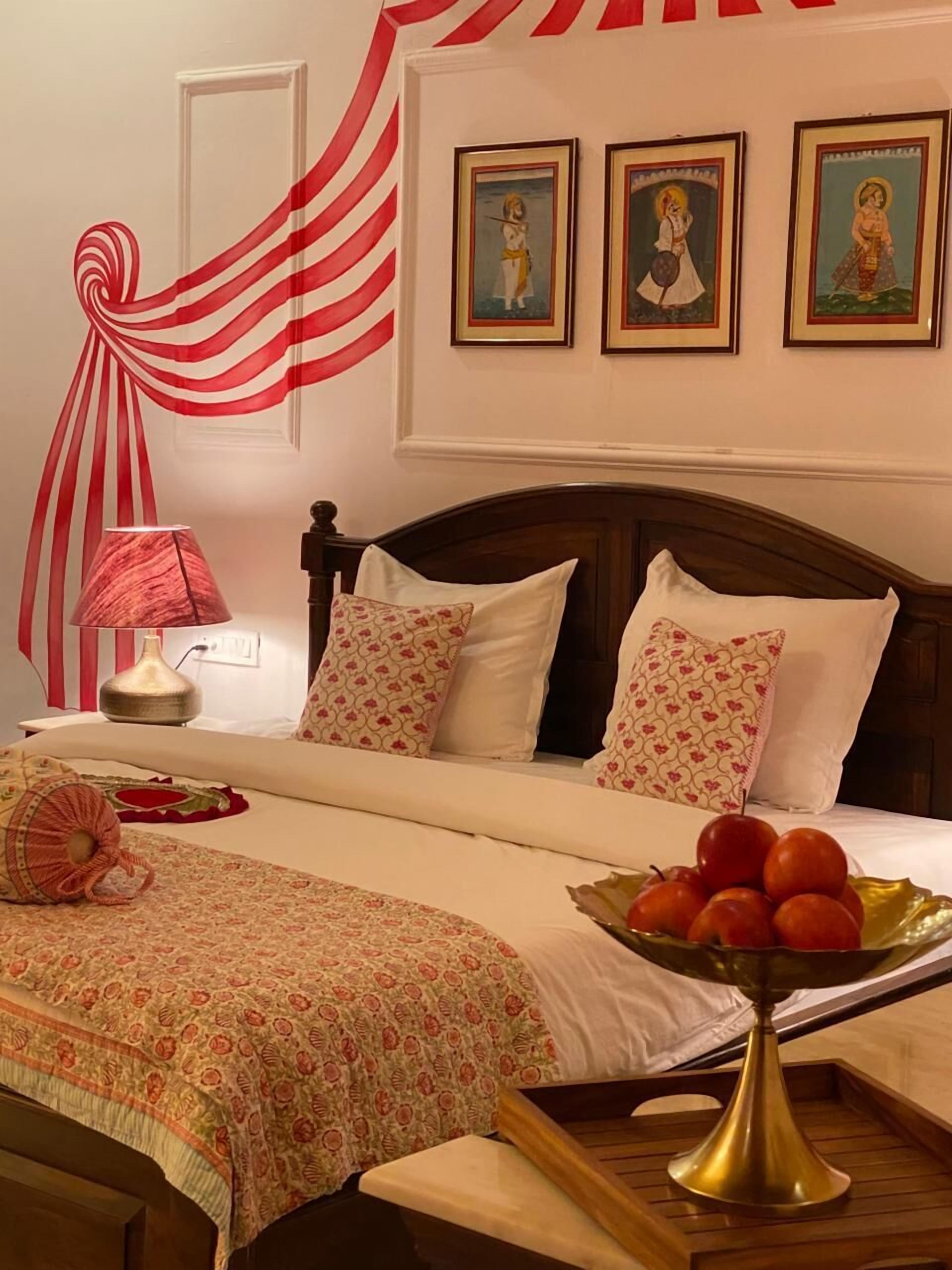 Une chambre d’hôtel avec un lit, une table de chevet avec une lampe, un vase décoratif rempli de fruits et trois tableaux encadrés accrochés au mur.