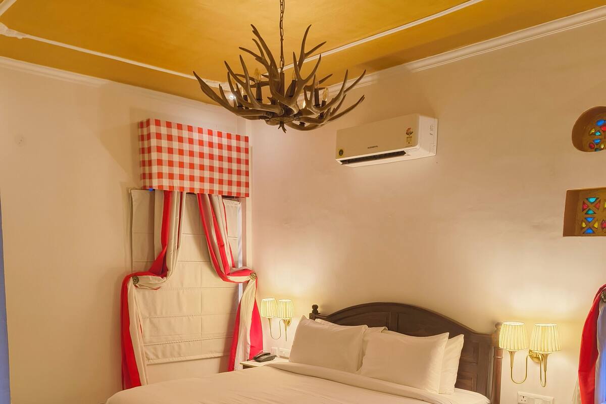 Un dormitorio con una cama de madera, un candelabro, una cortina a cuadros, una mesita de noche con lámparas y un aire acondicionado instalado en la pared.