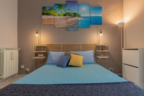 A bedroom with a bed, bedside tables, a nightstand, a wall-mounted lamp, a radiator, and a painting of a beach scene. - Oddone Comfy Apartment by Wonderful Italy (Torino)