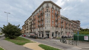 Un immeuble à plusieurs étages, avec une entrée située à l’angle, dont les façades sont composées d’un mélange de briques et de pierres, et qui présente de nombreux balcons ainsi qu’un espace commercial au rez-de-chaussée, avec une enseigne bien visible.