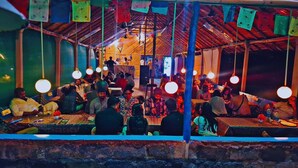 A crowded indoor space with people seated at tables, under a roof with hanging lights. - Shanthi Dhama Resort (Koppal)