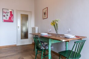 Un coin repas avec une table en bois dressée pour deux personnes, des chaises vertes, un vase avec des fleurs et une affiche accrochée au mur.