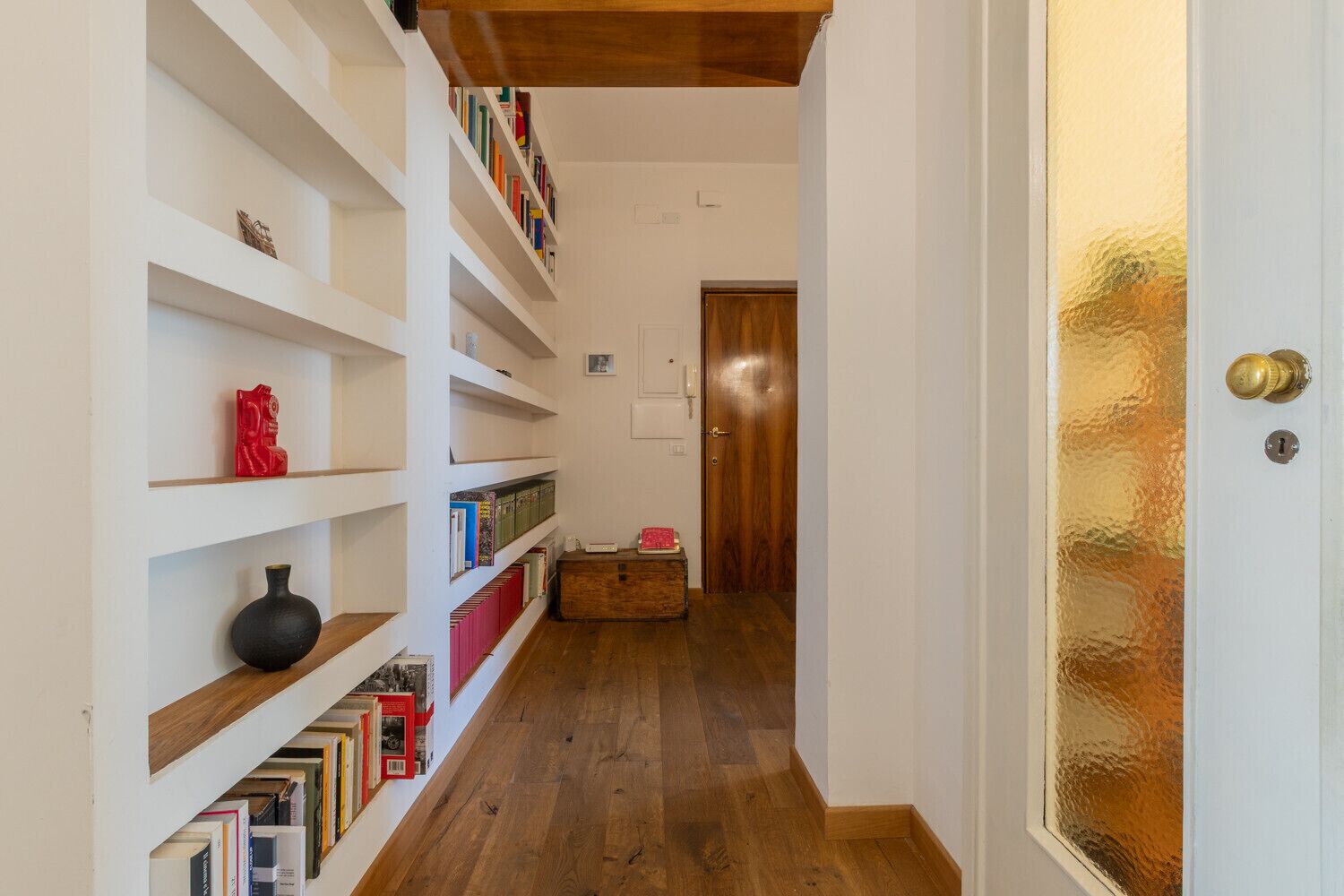 Passadís estret amb prestatgeries blanques plenes de llibres i objectes decoratius, que condueix a una porta de fusta amb un pom de bronze.