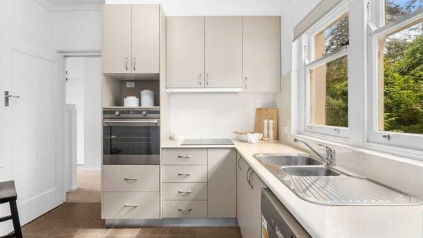 A modern kitchen with light-colored cabinets, a built-in oven, a dishwasher, and a window with a view of greenery. - Charming Cottage | Walk to City, Floriade & Eat Street (Braddon)