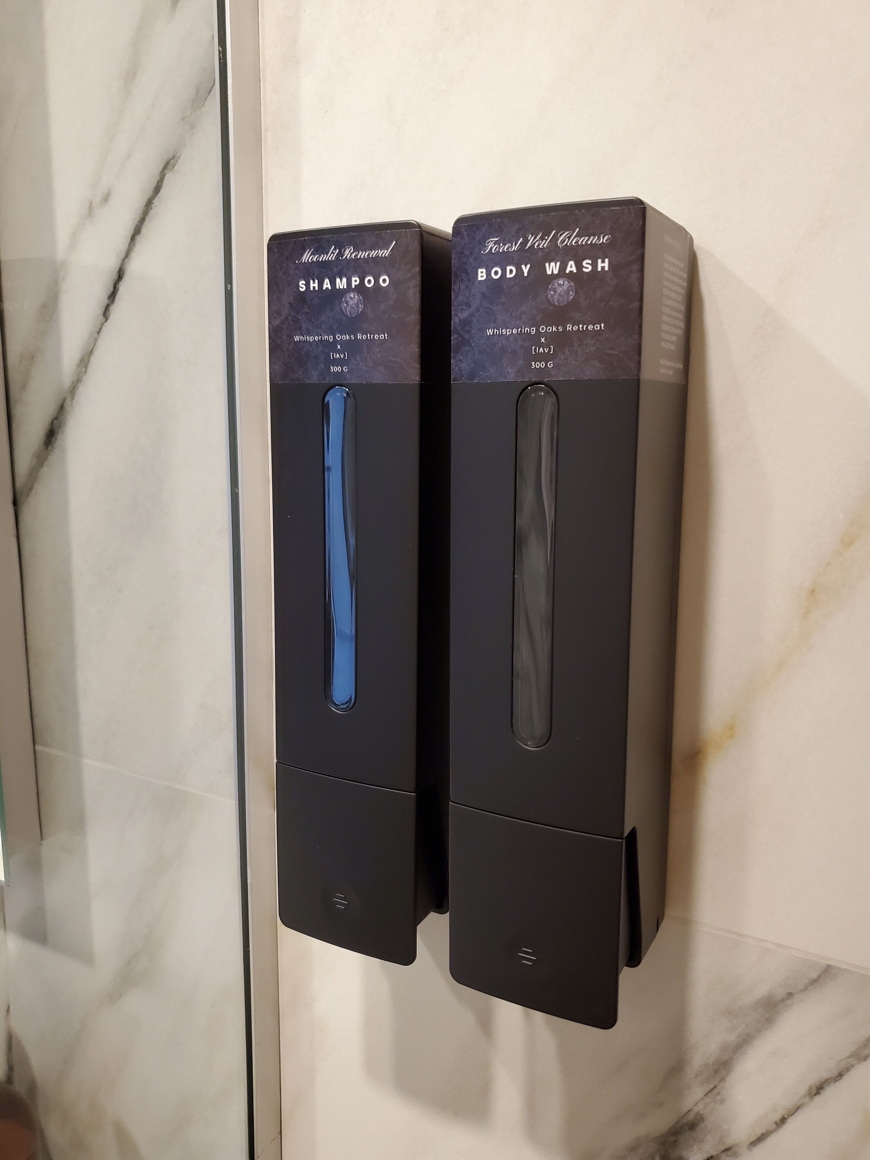 Two black dispensers for Moonlit Renewal shampoo and Forest Vail Essence body wash, each with a blue cap, mounted on a marble wall.