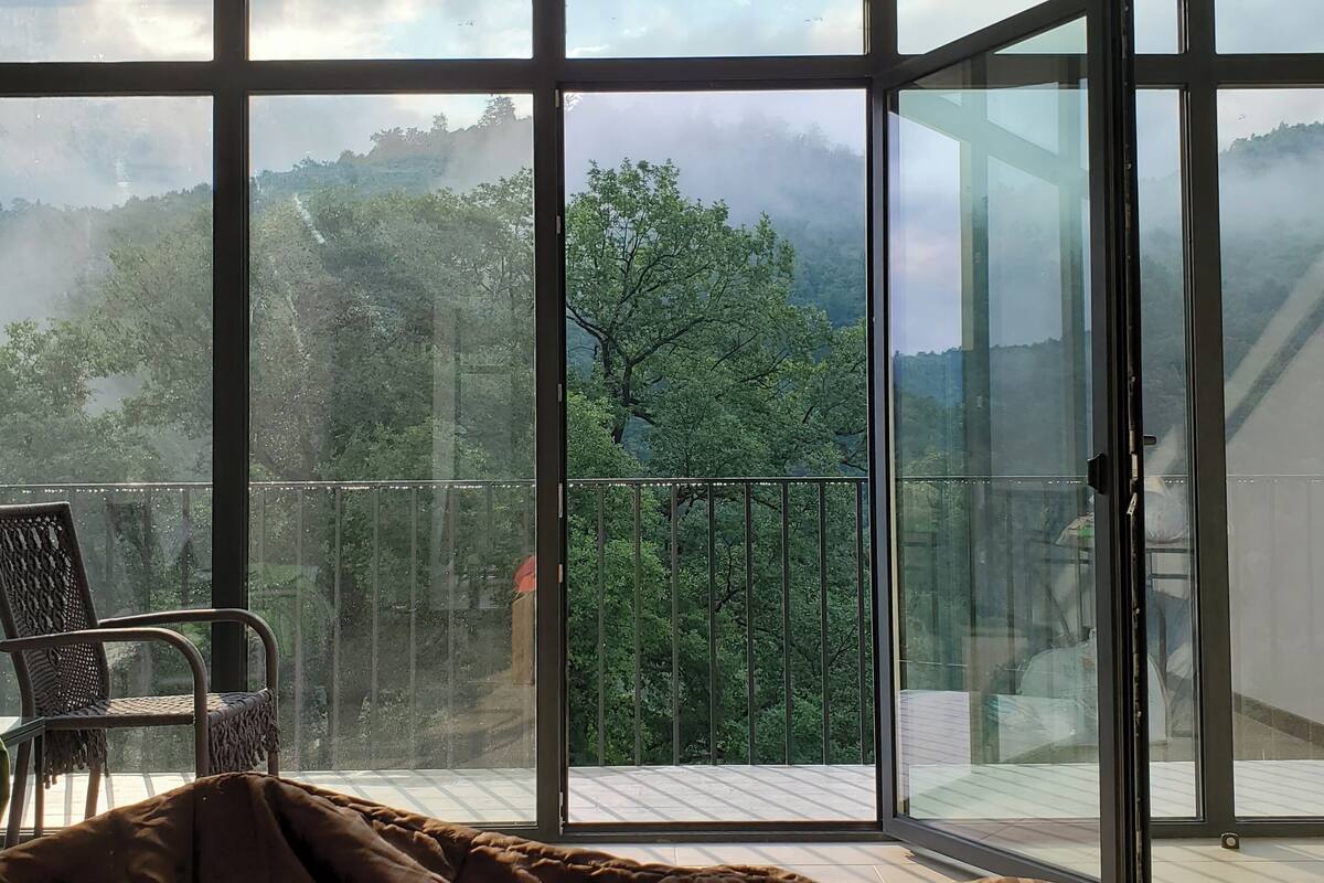 Un dormitorio con un amplio ventanal corredero que da a un balcón con vistas a una zona boscosa.