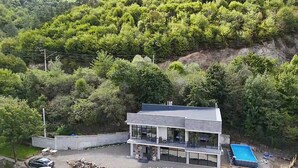 Una casa moderna de dos pisos con balcón, rodeada de vegetación exuberante y un pronunciado cerro.