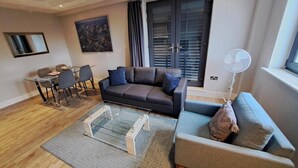 A modern living room with a dark grey sofa, a glass coffee table, and a grey sofa with a throw pillow. There is a dining area with a glass table and chairs, and a large window with blinds.