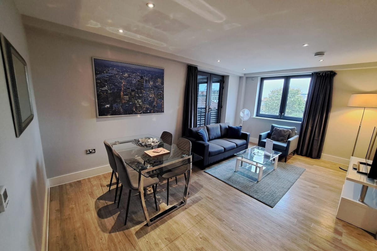 Een moderne woonkamer met een glazen eettafel, een bank en een salontafel. Er is een groot raam met gordijnen en uitzicht op de stad.