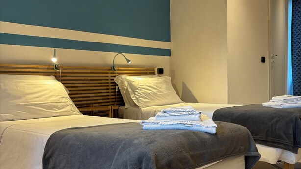 A hotel room with two beds, each with a wooden headboard and white bedding. There are two bedside lamps, a wall-mounted heater, and a door with a patterned curtain. - Dimora della Posta (Mondovì)
