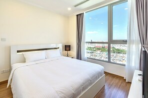 Habitación de hotel con una cama grande, una mesita de noche, una lámpara y vistas a la ciudad a través de amplios ventanales.