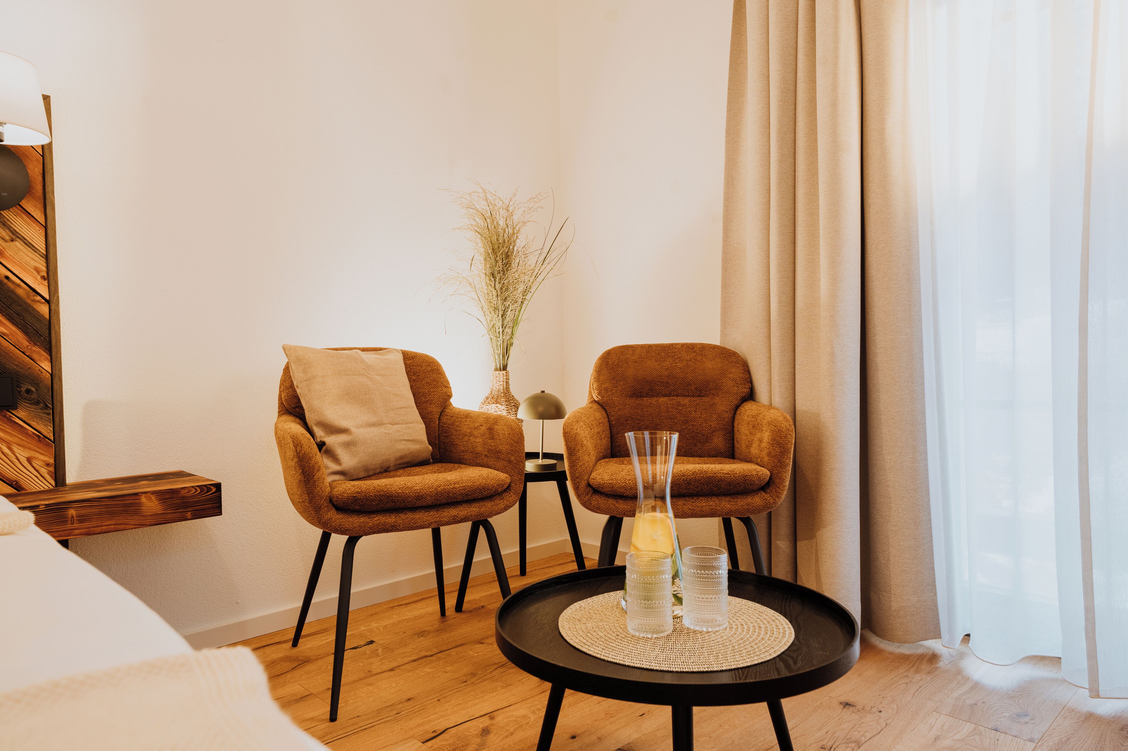 A room with two brown armchairs, a small table with a vase and glasses, a wooden sideboard, and a large window with sheer curtains.