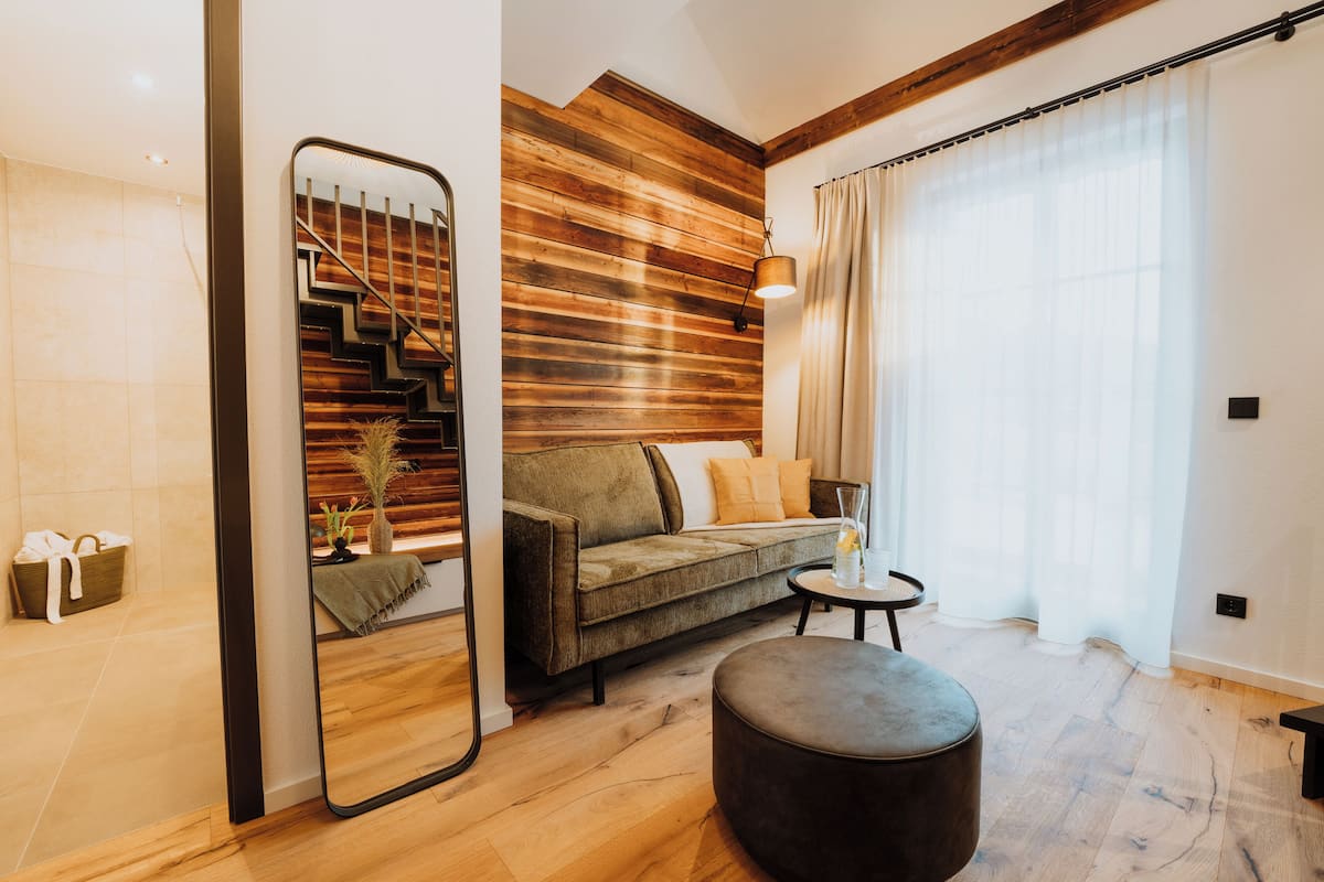 A modern living room with a wooden wall, a sofa, a round ottoman, and a mirror reflecting a staircase.