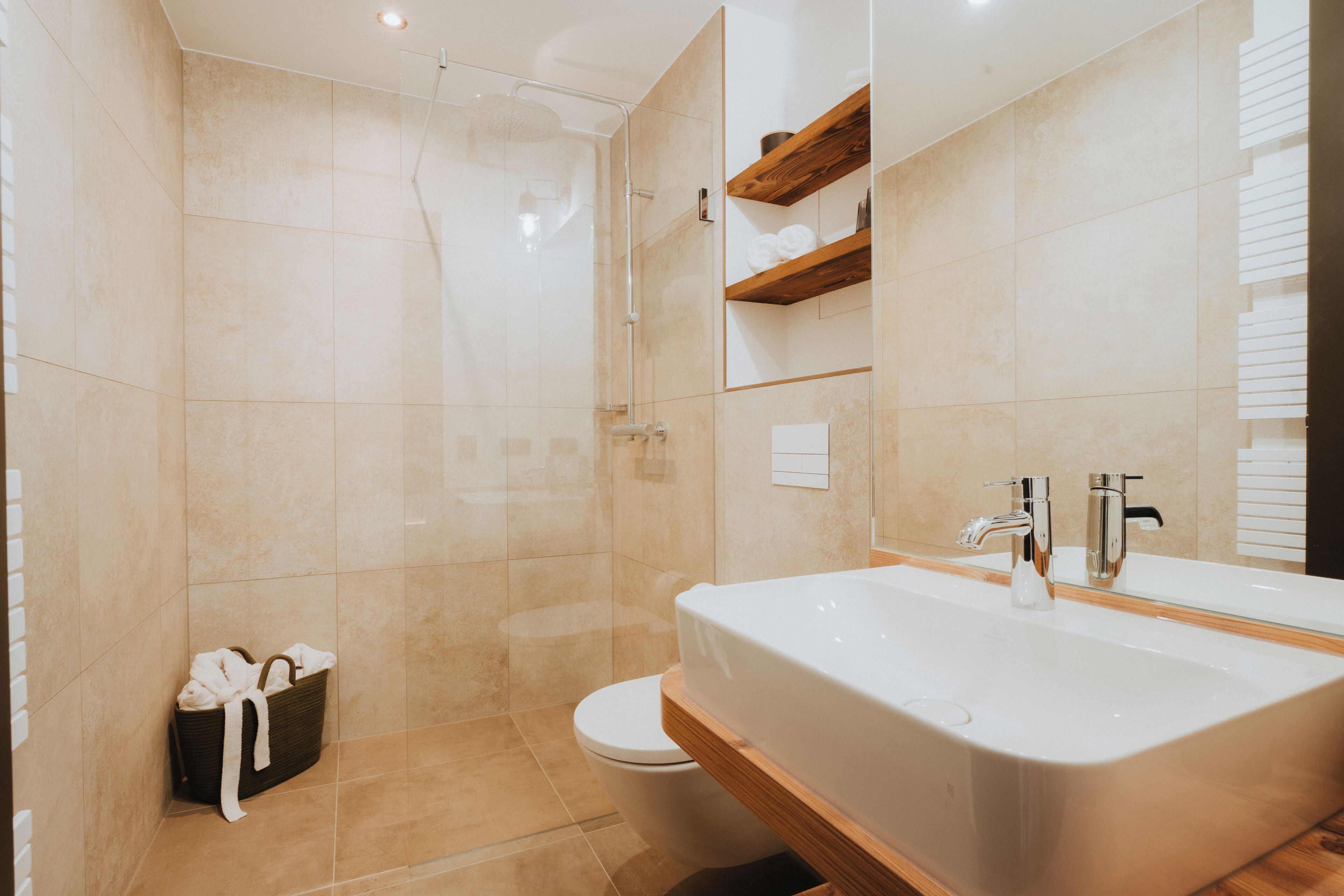 A modern bathroom with a large sink, a glass shower enclosure, and a wooden shelf above the toilet.