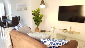 Una sala de estar con un sofá marrón, cojines estampados, una mesa de centro de vidrio, una lámpara de pie, un televisor y un comedor con sillas y una mesa.