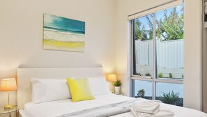 Habitación de hotel con cama, un cuadro de una playa, ventana con vistas a palmeras y una lámpara de noche.