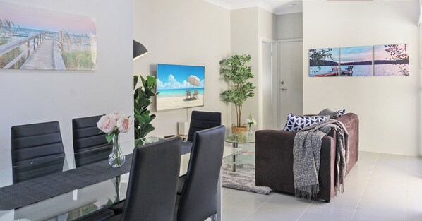 A modern living room with a glass dining table, black chairs, a brown sofa, and a large window.