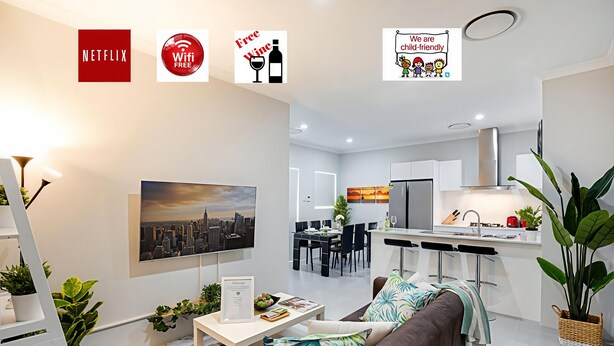 A modern living room with a brown sofa, a coffee table, and a cityscape painting. There is a dining area with a table and chairs, and a kitchen with white cabinets and a stainless steel range hood. - Culloton Escape – Spacious 3BR Family Home Near Parks & Shops (Balga)
