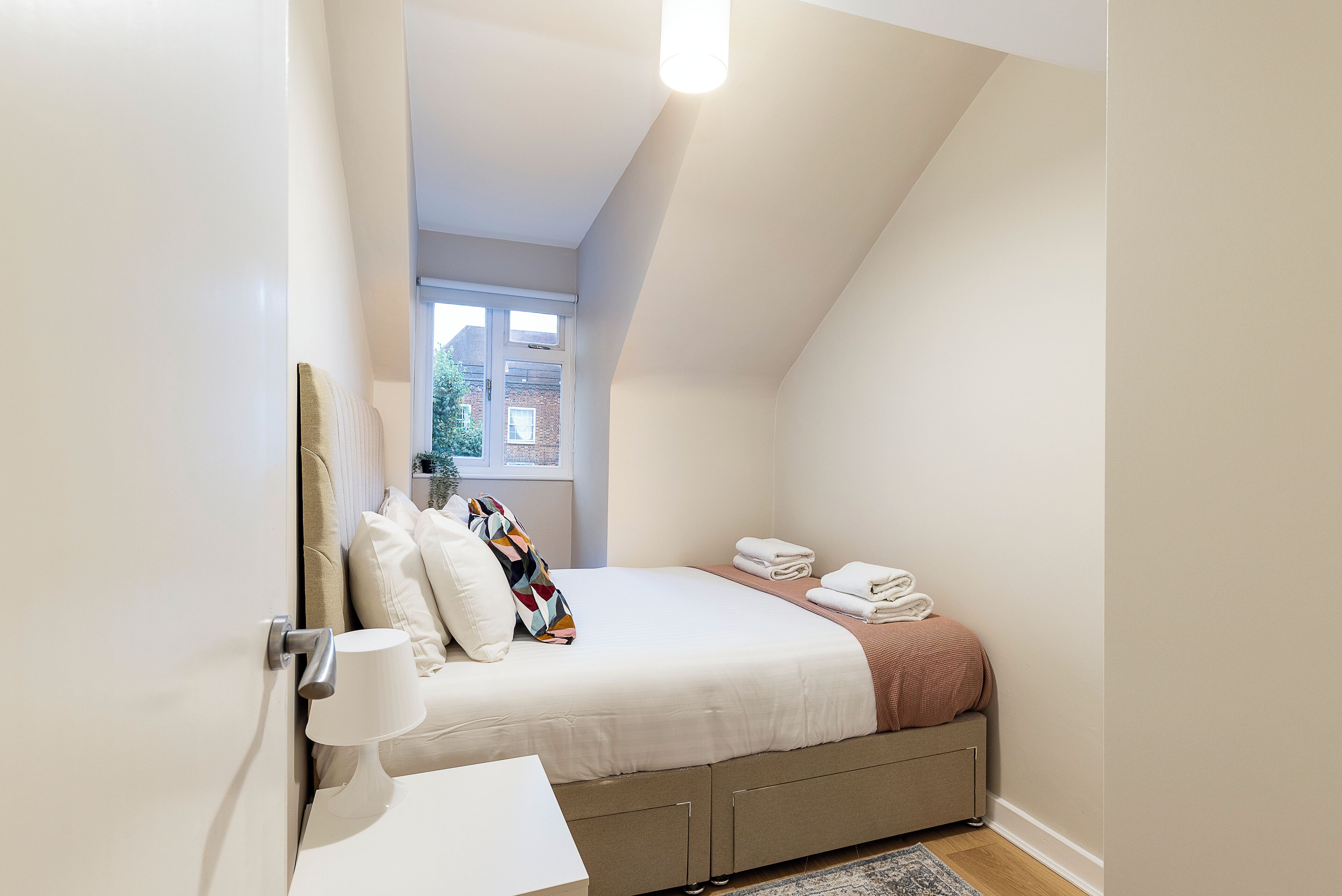 A bedroom with a bed, pillows, a bedside lamp, a window, and a view of the outside.