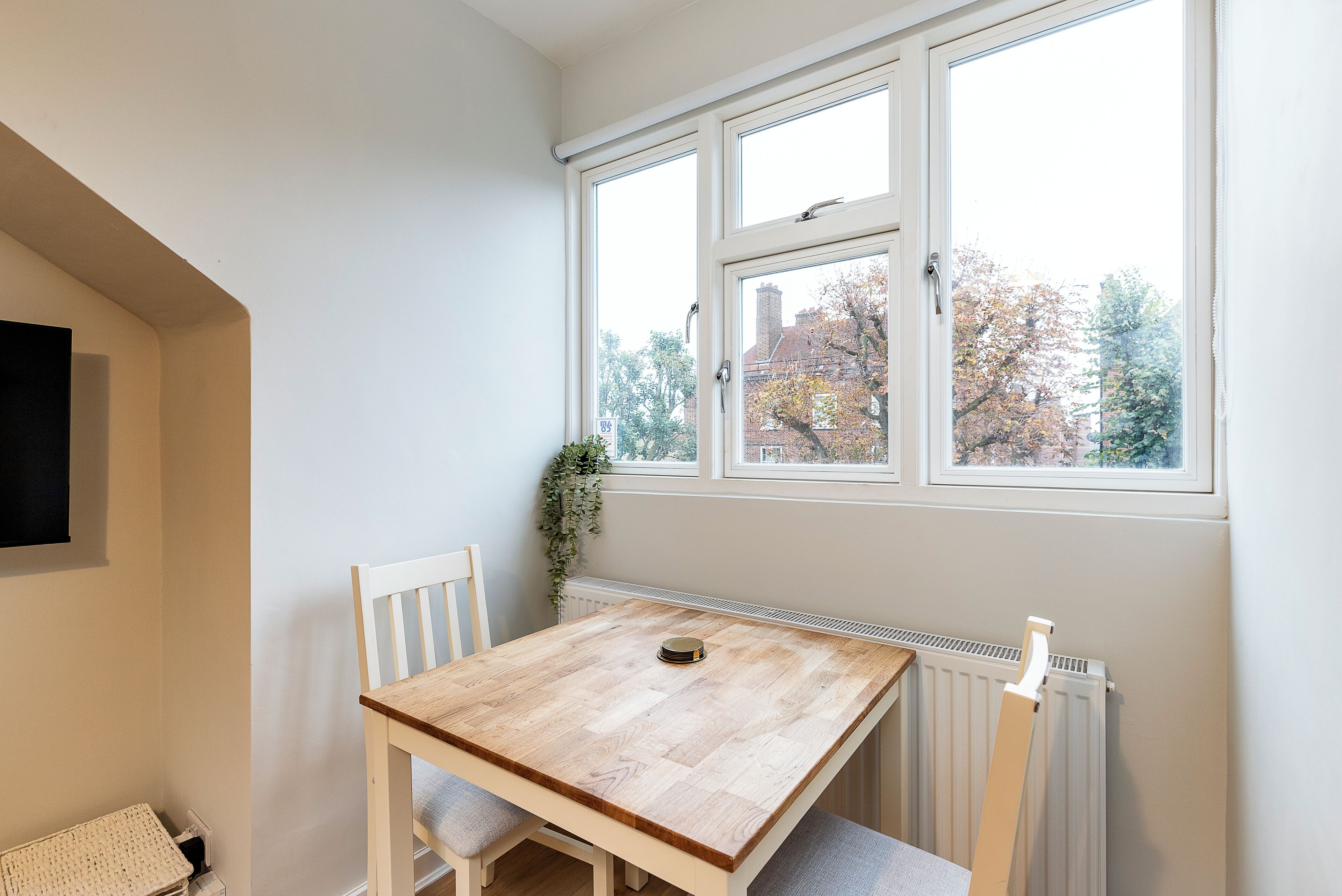 Een kamer met een houten tafel en twee stoelen, een radiator en een raam met uitzicht op bomen en gebouwen.