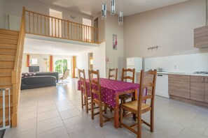 Un coin repas avec une table dressée pour quatre personnes, des chaises en bois, une cuisine équipée d’appareils électroménagers modernes et un escalier menant à l’étage supérieur.