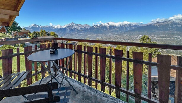 Un balcon avec une table et des chaises, donnant sur un paysage montagneux aux sommets enneigés et sous un ciel bleu dégagé.