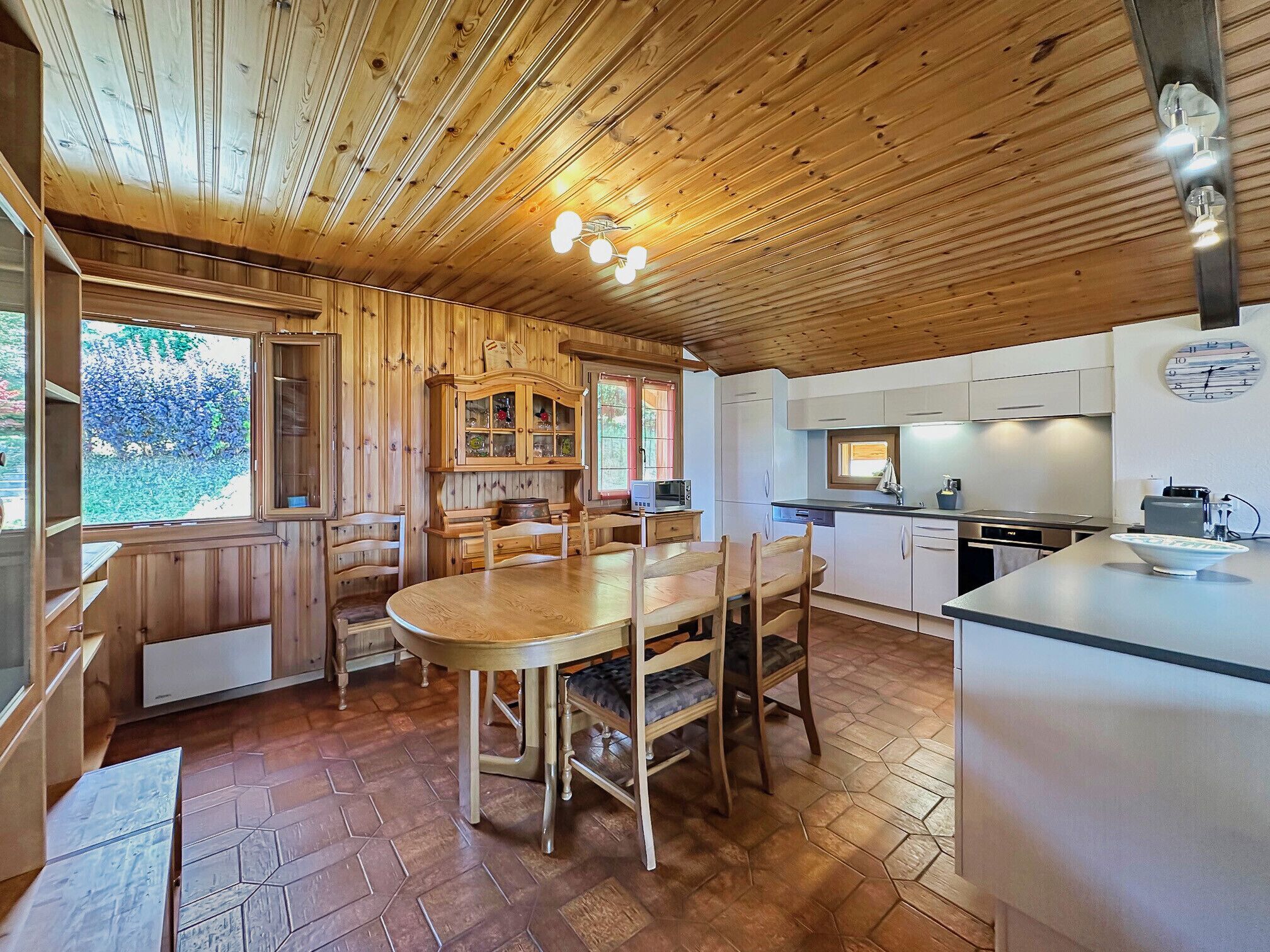 A wooden interior with a dining table, chairs, a kitchen area, and a clock on the wall.
