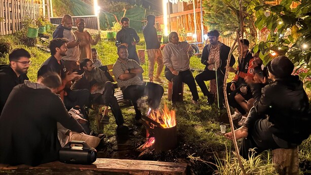 Un grupo de personas reunidas alrededor de una fogata en un entorno al aire libre, con construcciones de madera y vegetación de fondo.