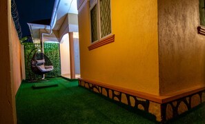 Un sendero estrecho que conduce a un edificio con fachada amarilla y una ventana con persiana de diseño geométrico.