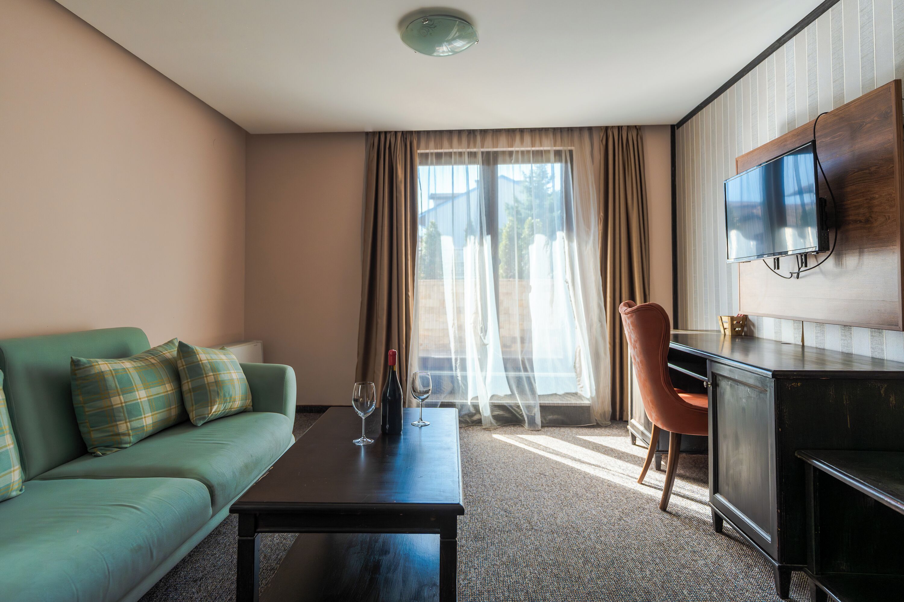 Une chambre d’hôtel avec un canapé vert, un bureau en bois, un téléviseur fixé au mur et une fenêtre ornée de rideaux légers.